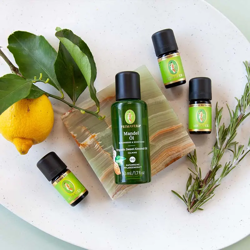 A white bowl with a lemon and green leaves, PRIMAVERA, and three bottles of essential oils— Peppermint org., Rosemary Verbenon org.  Lemon org. a light blue background.