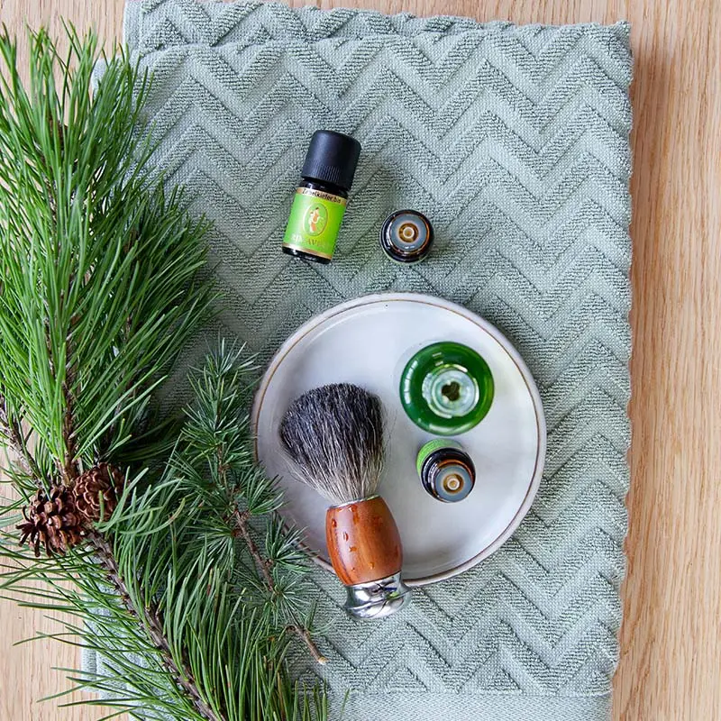A shaving brush, Swiss Pine org., two additional open essential oil bottles, a bottle of organic skin care oil seen from above, and pine branches decoratively arranged on a green towel against a wooden background.