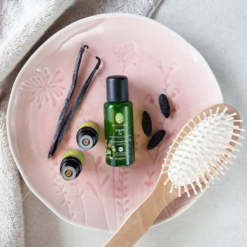 A pink plate with a floral pattern, on which are three green oil bottles; below them are organic tonka extract, Vanilla Extract org. , and Organic Argan Seed Oil; two vanilla beans, three tonka beans, a brush, and a beige towel in the background.