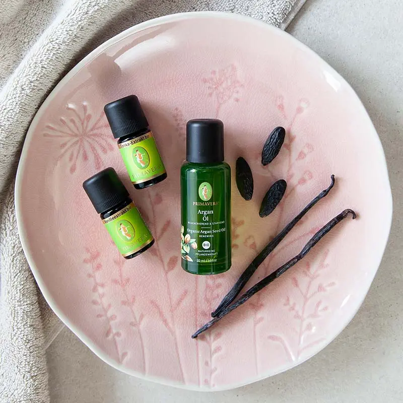 A pink plate with a floral pattern, with three green oil bottles on it, including organic tonka extract, Vanilla Extract org. , and Organic Argan Seed Oil; two vanilla beans, three tonka beans, and a beige towel in the background.