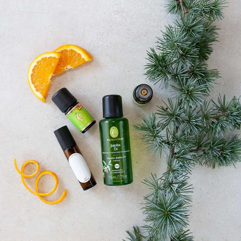Two bottles of essential oil, including Cedar Wood org. and Orange demeter , as well as Organic Plant Oil Organic Jojoba Oil . Surrounding them is a Roll-On bottle with a blank label, orange slices, and pine twigs.