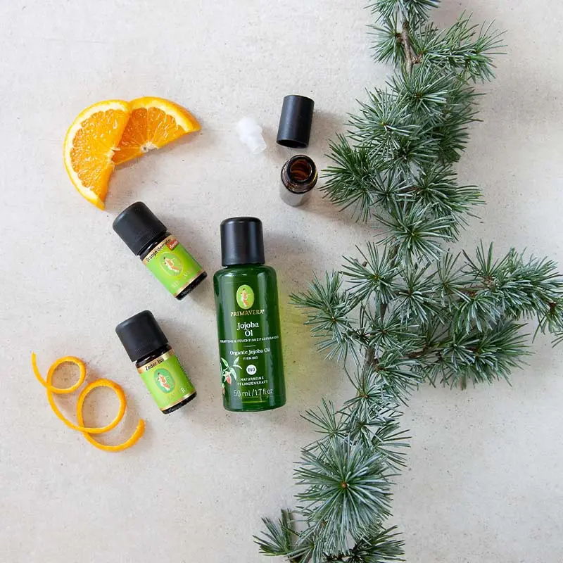 Two small bottles of essential oil, including " Cedar Wood org. " and "Orange" demeter , as well as " Organic Plant Oil  "Organic Jojoba Oil. Arranged around them are an empty Roll-On bottle, orange slices, and pine twigs.