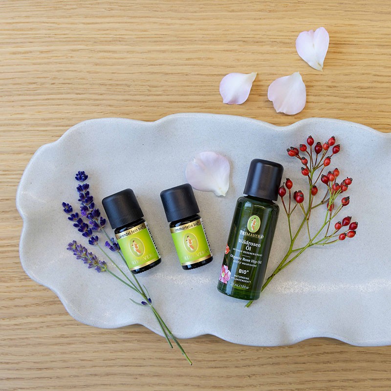 Wildrosenöl bio, Lavendel fein bio und Immortelle bio auf einer hellen Schale, dekoriert mit Lavendelblüten, einer Rosenknospe und Hagebutten.