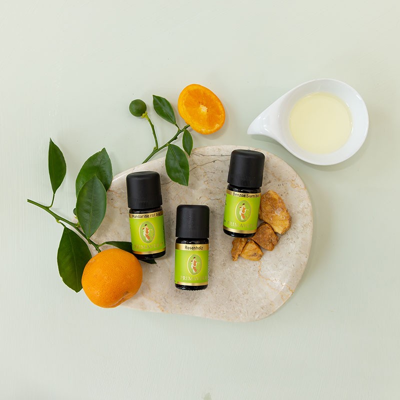 Ätherischer Öle Benzoe Siam, Mandarine und Rosenholz auf einer Steinschale, daneben ein Schälchen Körperöl, dekorativ arrangiert mit frischen Mandarinenzweigen.