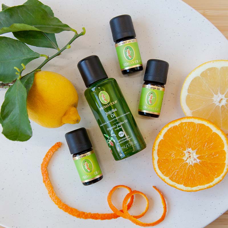 Weißer Teller mit Zitrone, Zitrusscheiben, Jojobaöl bio, Orangenöl bio, Grapefruitöl bio und Zitrone bio auf Holzuntergrund.