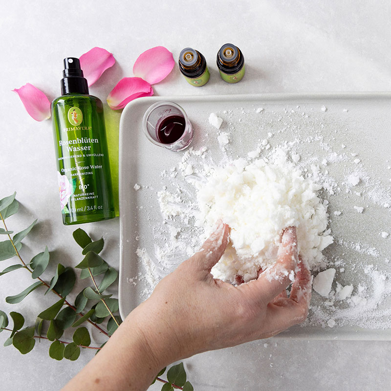 DIY Badepralinen-Zubereitung: Hände formen Mischung aus Naturkosmetik-Zutaten, daneben Primavera Rosenblütenwasser, ätherische Öle und Rosenblätter.