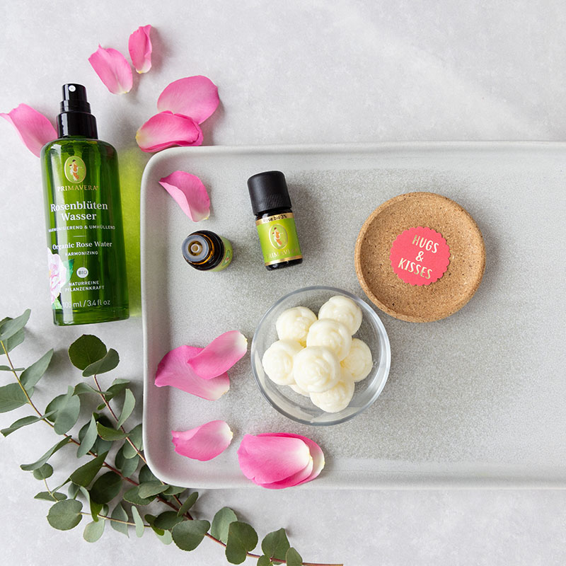 Primavera Rosenblütenwasser, ätherische Öle und weiße Badepralinen auf rosa Teller mit Rosenblättern dekoriert.