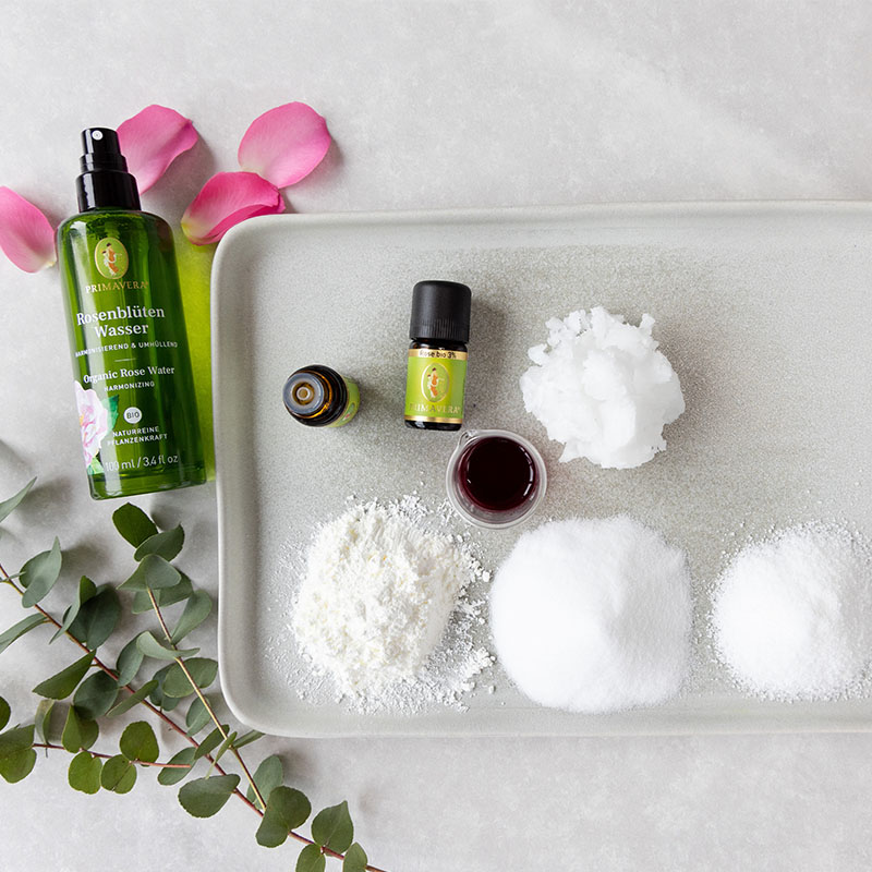 DIY Badepralinen-Zutaten mit Primavera Rosenblütenwasser, ätherischen Ölen und Naturkosmetik-Zutaten auf Tablett, dekoriert mit Rosenblättern und Eukalyptus.