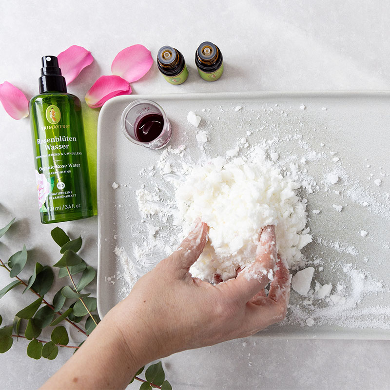 DIY Badepralinen-Zubereitung: Hände formen Mischung aus Naturkosmetik-Zutaten, daneben Primavera Rosenblütenwasser, ätherische Öle und Rosenblätter.