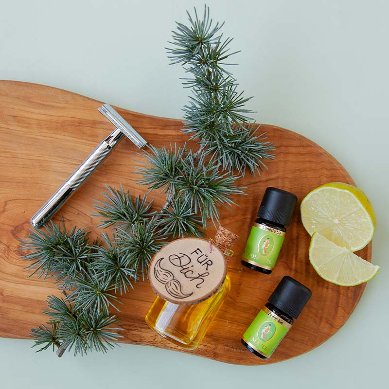 Holzbrett mit Rasierer, Kiefernzweigen, Fläschchen mit Holzanhänger und der Beschrifung: „Für Dich“, Ätherische Öle Limette bio und Zeder bio und zwei Limettenscheiben.