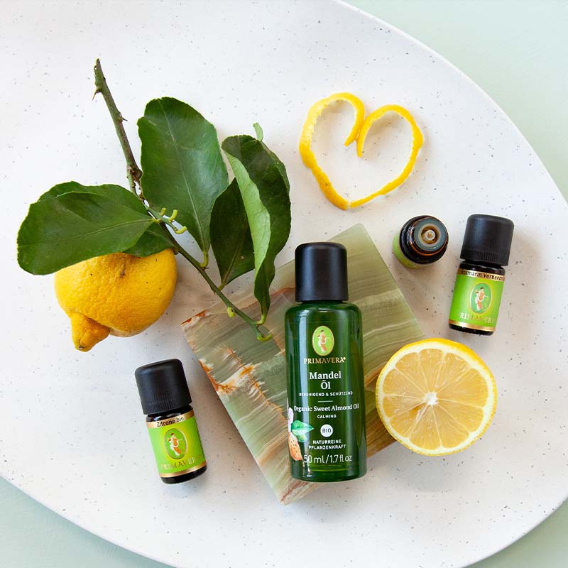 Weiße Schale mit Zitrone und grünen Blättern, PRIMAVERA-Mandelöl, drei ätherischen Ölflaschen, Pfefferminze bio, Rosmarin Verbenon bio und Zitrone bio auf einer hellblauen Unterlage.
