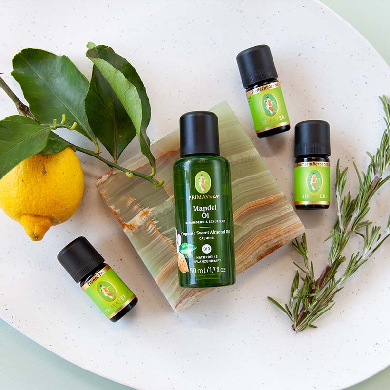 Weiße Schale mit Zitrone und grünen Blättern, PRIMAVERA-Mandelöl, drei ätherischen Ölflaschen, Pfefferminze bio, Rosmarin Verbenon bio und Zitrone bio auf einer auf einer hellblauen Unterlage.