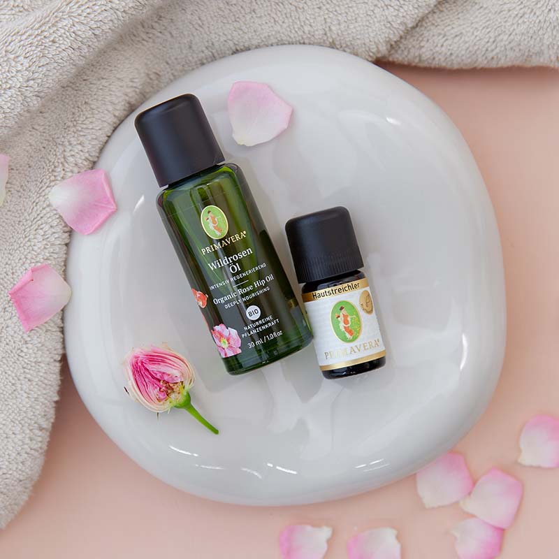 PRIMAVERA Wildrosenöl und Hautstreichler mit Rosenblüten auf weißem Untergrund, umgeben von Blütenblättern und beiger Decke.