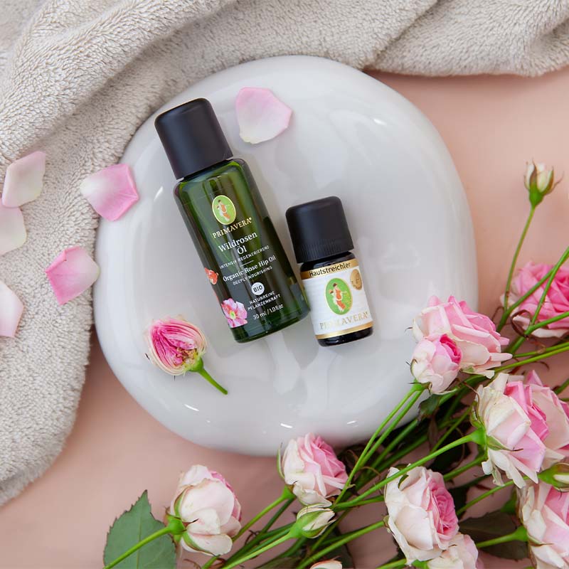PRIMAVERA Wildrosenöl und Hautstreichler mit Rosenblüten auf weißem Untergrund, umgeben von Blütenblättern und beiger Decke.