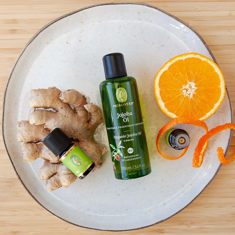 Weißer Teller mit Jojobaöl, Ingwer bio und Orange bio, Ingwerstück und zwei Orangenscheiben auf Holzuntergrund.