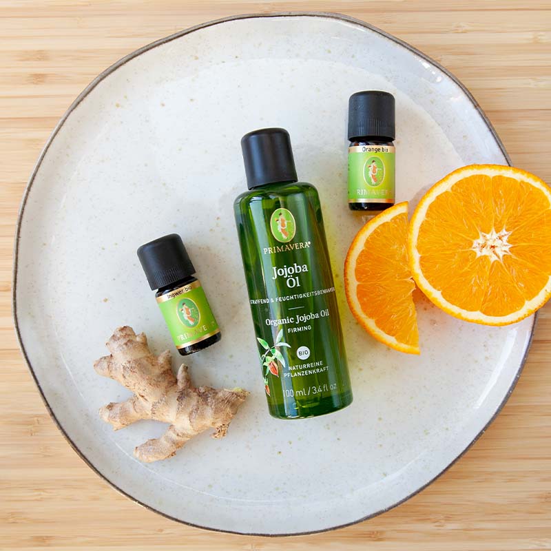 Weißer Teller mit Jojobaöl, Ingwer bio und Orange bio, Ingwerstück und zwei Orangenscheiben auf Holzuntergrund.