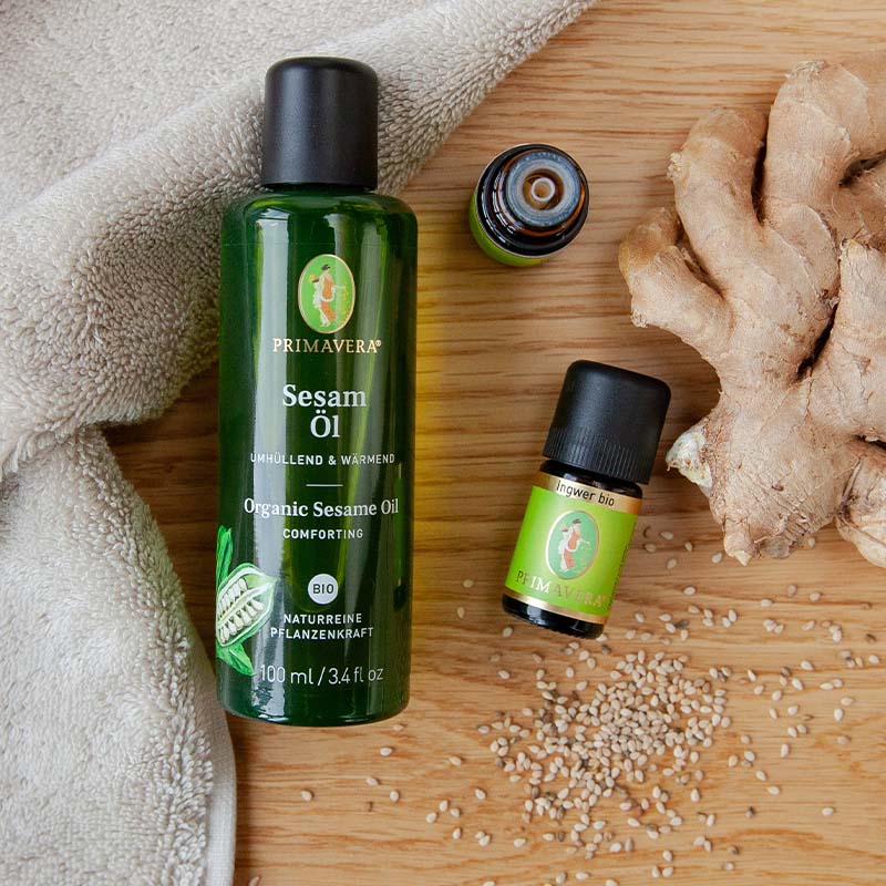 PRIMAVERA-Sesamöl, Ingweröl, Sesamsamen, Ingwerstück und beiges Handtuch auf Holzuntergrund.