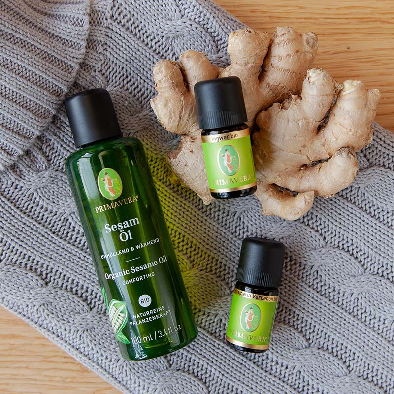 Graue Wärmflasche, auf dieser liegen das PRIMAVERA-Sesamöl, Ätherischen Öle Ingwer bio und Rosmarin Verbenon bio und frischer Ingwer, auf einem Holzuntergrund.