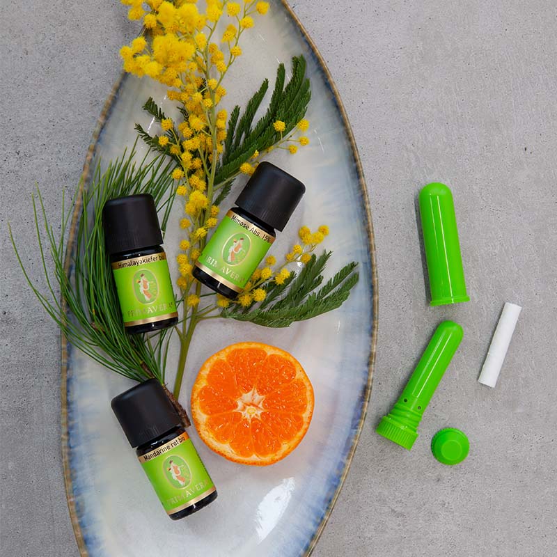 Drei Primavera-Ölfläschchen mit Blüten, Blättern und halber Orange auf Teller, daneben zwei Riechsticks.