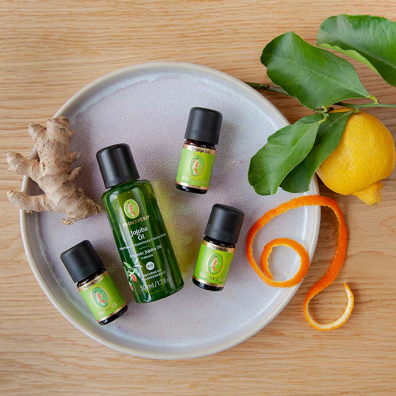 Teller mit ätherischen Ölfläschchen Zitrone bio, Orange bio, Ingwer bio, Ingwer, Zitrone mit Blättern und Orangenschale in Spirale auf Holzuntergrund.