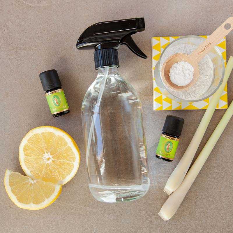 Sprayflasche mit den ätherischen Ölen Grapefruit bio und Lemongrass bio, halber Zitrone, Zitronengras, Schale mit Natron und Holzlöffel auf grauem Untergrund.