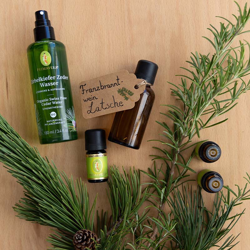 Ein braunes Fläschchen auf hellem Holz mit Ettikett „Franzbranntwein Latsche“. Daneben liegt ein ätherisches Öl Fläschen Latschenkiefer bio 5 ml, Zirbelkiefer Zedernwasser, 2 offene ätherische Öl Fläschchen und grüne Zweige.
