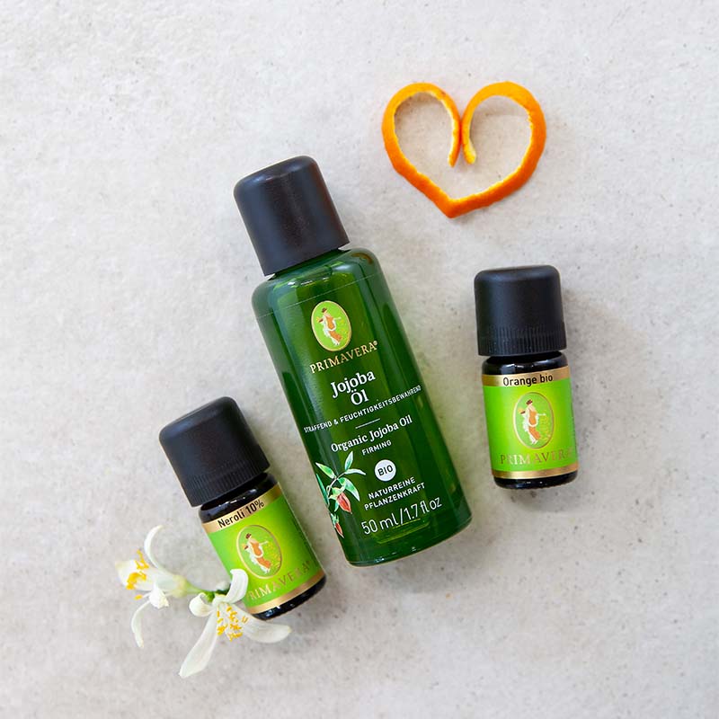 Orangenschale in Form von einem Herz, PRIMAVERA-Jojobaöl, PRIMAVERA Ätherische Öle Orange bio und Neroli 10 % und zwei Orangenblüten auf hellem Untergrund.