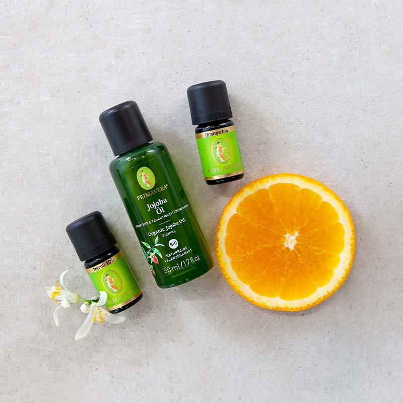 Orangenscheibe, PRIMAVERA-Jojobaöl, PRIMAVERA Ätherische Öle Orange bio und Neroli 10 % und zwei Orangenblüten auf hellem Untergrund.