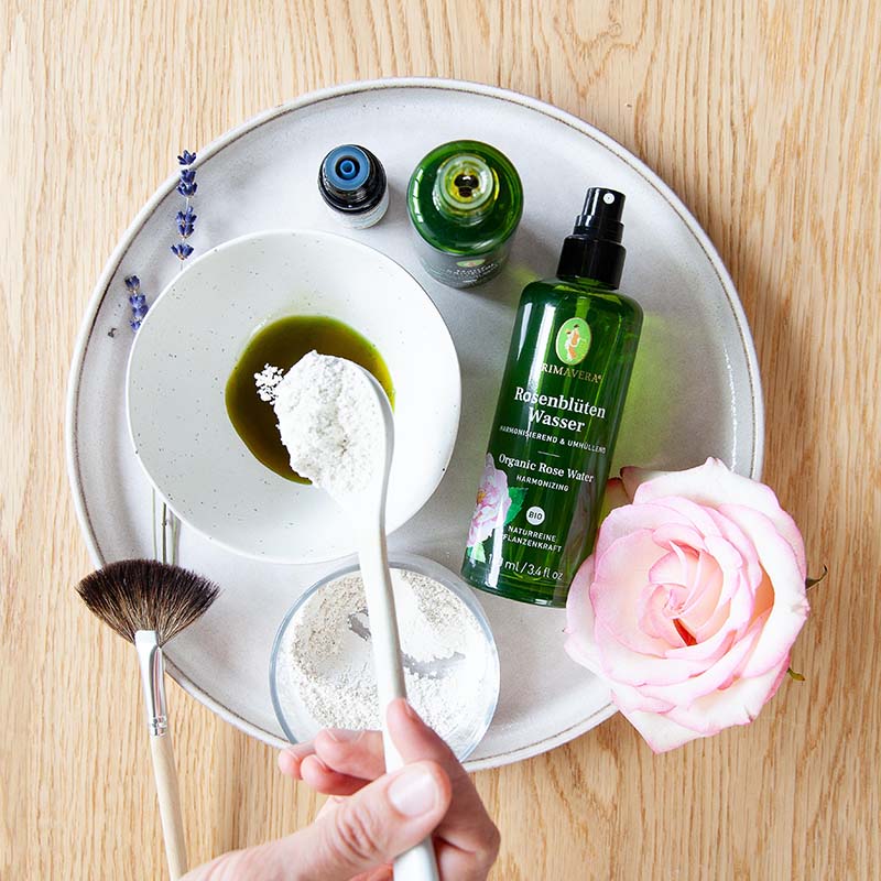 Runde Schale mit Rosenblütenwasser, Schale mit weißer Tonerde und ein weiteres Gefäß mit Hanfsamenöl, Löffel, Pinsel, Lavendel und rosa Rose.