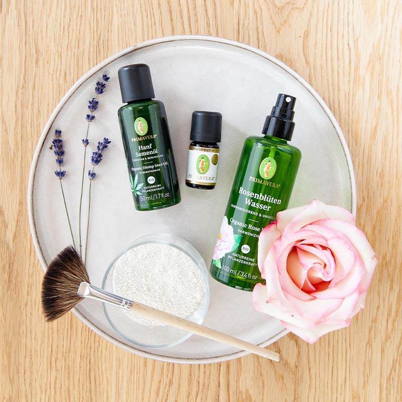 Runde Platte mit PRIMAVERA Hanfsamenöl, Rosenblütenwasser, Hautstreichler Öl, Lavendel, weiße Tonerde in extra Gefäß, Pinsel und rosa Rose auf Holzuntergrund.