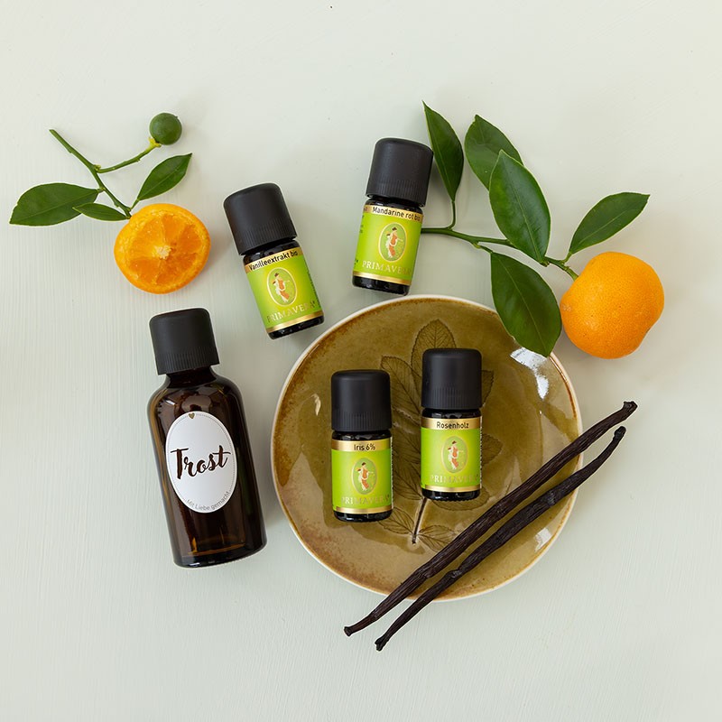 Die ätherischer Öle Vanilleextrakt, Mandarine rot, Iris 6% und Rosenholz mit einer braune Flasche mit der Aufschrift ‚Trost‘, dekorativ arrangiert mit frischen Mandarinenzweigen und Vanilleschoten auf hellem Untergrund