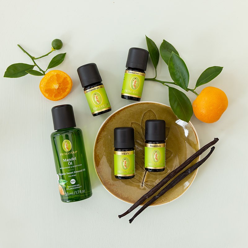 Die ätherischer Öle Vanilleextrakt, Mandarine rot, Iris 6% und Rosenholz mit PRIMAVERA Mandelöl dekorativ arrangiert mit frischen Mandarinenzweigen und Vanilleschoten auf hellem Untergrund