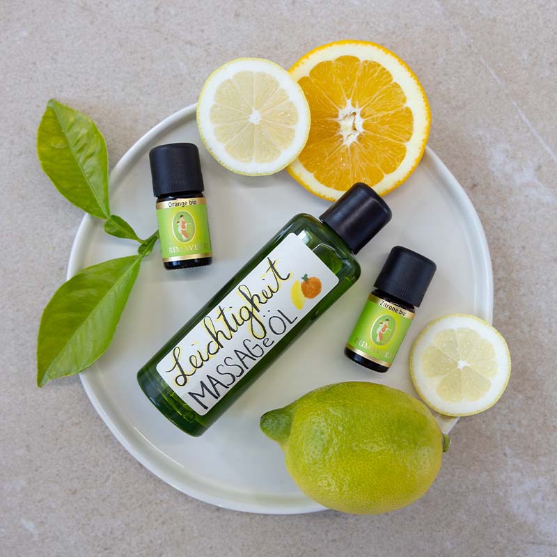 Zutaten fürs DIY-Rezept: Ätherische Öle Orange bio und Zitrone bio, Jojobaöl bio und Etikett „Leichtigkeit Massageöl“. Im Hintergrund befinden sich Zitrusfrüchtescheiben und grüne Blätter.