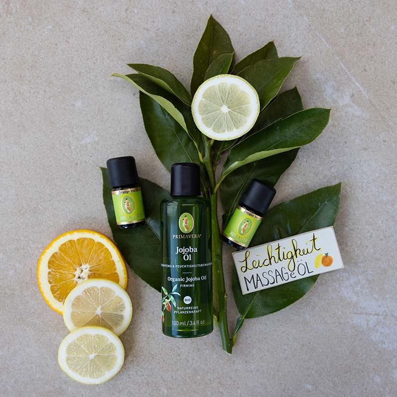 Zutaten fürs DIY-Rezept: Ätherische Öle Orange bio und Zitrone bio, Jojobaöl bio und Etikett „Leichtigkeit Massageöl“. Im Hintergrund befinden sich Zitrusfrüchtescheiben und grüne Blätter.