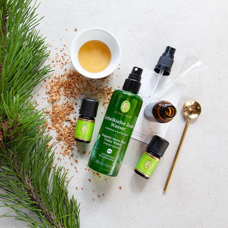 Zirbelkiefer Zeder Wasser, Ätherische Öle Orange bio und Propois Extrakt Allgäu, Pipettenflasche, Schale mit Hanfsamenöl bio, goldener Löffel, Kiefernzweige und Propolis, leere Sprühflasche mit Etiketten auf grauem Untergrund.