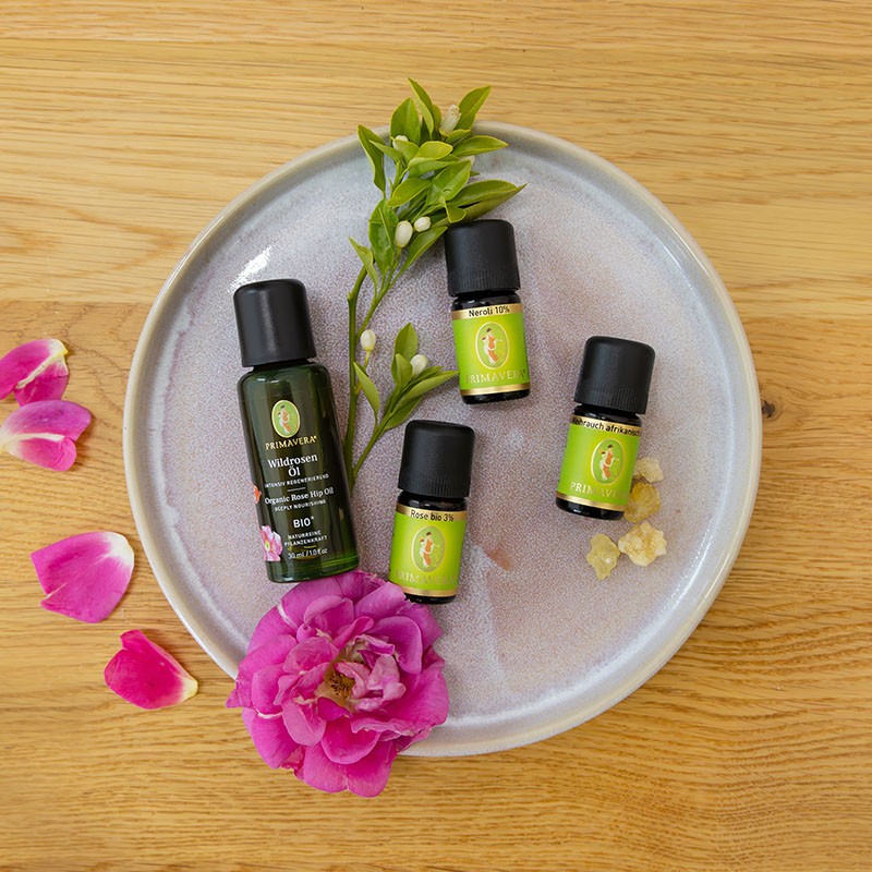 PRIMAVERA Wildrosen Öl mit ätherischen ölen Neroli 10%, Weihrauch afrikanisch und Rose bio 3% auf rundem Teller mit Rosen Blüten und grünen Blättern auf einem Holz Hintergrund.