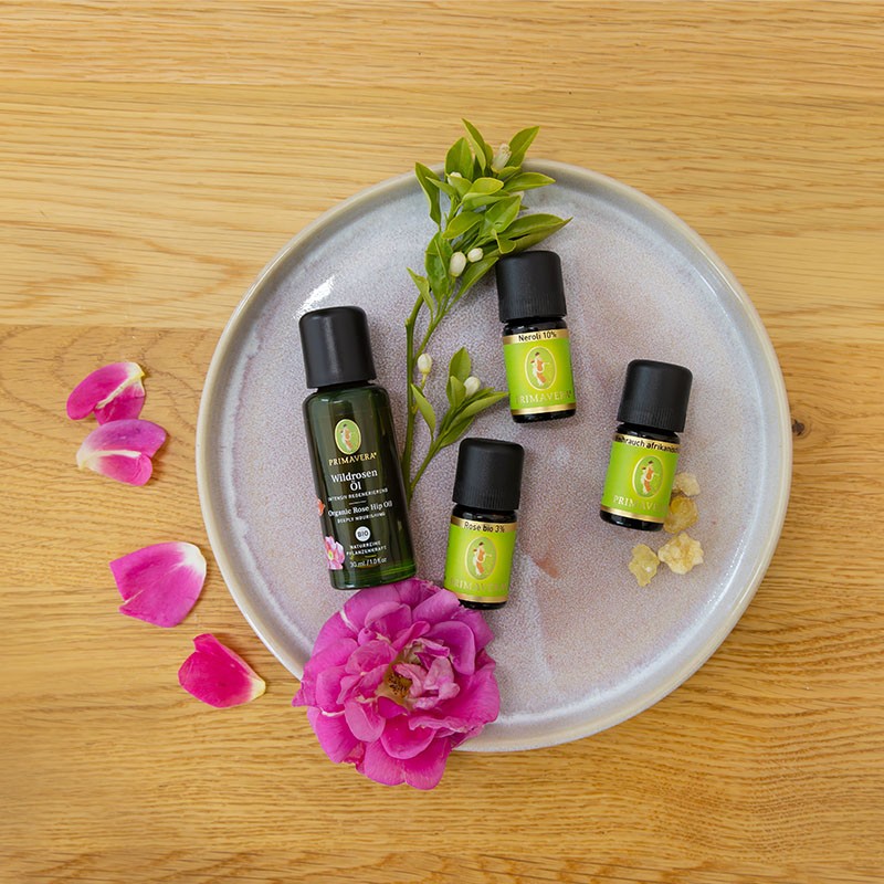 PRIMAVERA Wildrosen Öl mit ätherischen ölen Neroli 10%, Weihrauch afrikanisch und Rose bio 3% auf rundem Teller mit Rosen Blüten und grünen Blättern auf einem Holz Hintergrund.