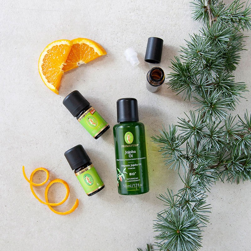 Zwei ätherische Ölfläschchen, darunter Zeder bio und Orange demeter und das Bio Pflegeöl Jojobaöl bio. Drumherum befindet sich eine Roll-On Flasche mit einem leeren Etikett, Orangenscheiben und Kiefernzweige.