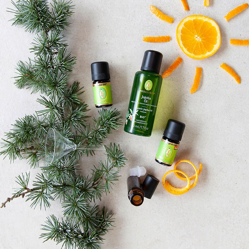 Zwei ätherische Ölfläschchen, darunter Zeder bio und Orange demeter und das Bio Pflegeöl Jojobaöl bio. Drumherum befindet sich eine leere Roll-On Flasche, Orangenscheiben und Kiefernzweige.