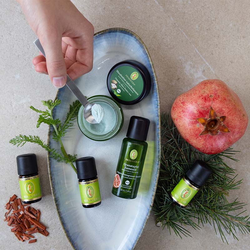 Ätherische Öle des Rezepts, Sheabutter und Granatapfelsamenöl auf einer Schale mit Granatapfel und einem Nadelzweig dekorativ arrangiert. Zudem wird mit einem Löffel ein Teil des fertigen Balsams entnommen.