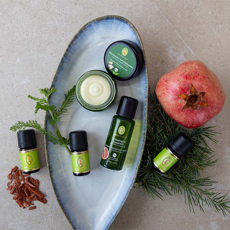 Ätherische Öle des Rezepts, Sheabutter und Granatapfelsamenöl auf einer Schale mit Granatapfel und einem Nadelzweig dekorativ arrangiert.