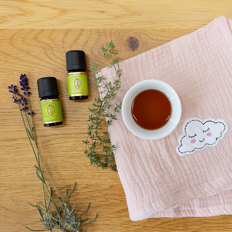 Die ätherischen öle Lavendel fein bio und Thymian Linalool bio, von PRIMAVERA, mit einer Schale des hergestellten Körperöls auf rosa Tuch auf Holzuntergrund.