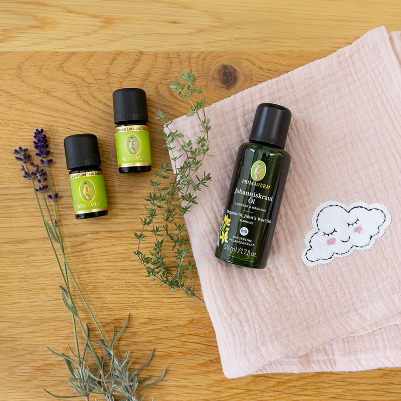 PRIMAVERA Johanniskrautöl mit ätherischen ölen Lavendel fein bio und Thymian Linalool bio, dekoriert mit frischen Kräutern und rosa Tuch auf Holzuntergrund.