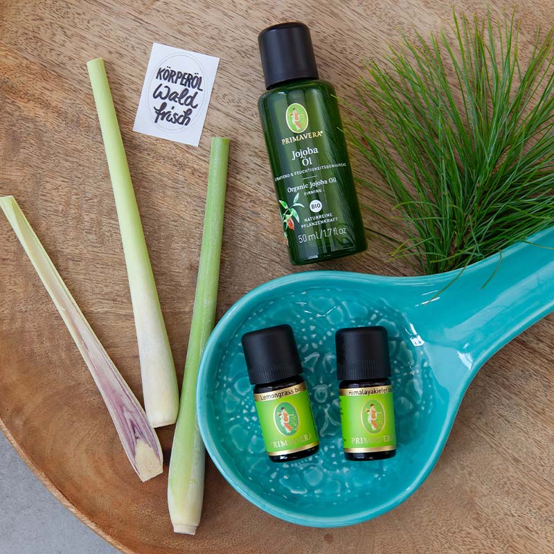 Auf einer Holzplatte liegen in einem blauen größeren Löffel die beiden Ätherischen Öle, Lemongrass bio und Himalayakiefer bio, darüber das Jojobaöl. Links davon ist ein halbierter Zitronengrasstängel und rechts zwei Zweige mit Kieferngrün.